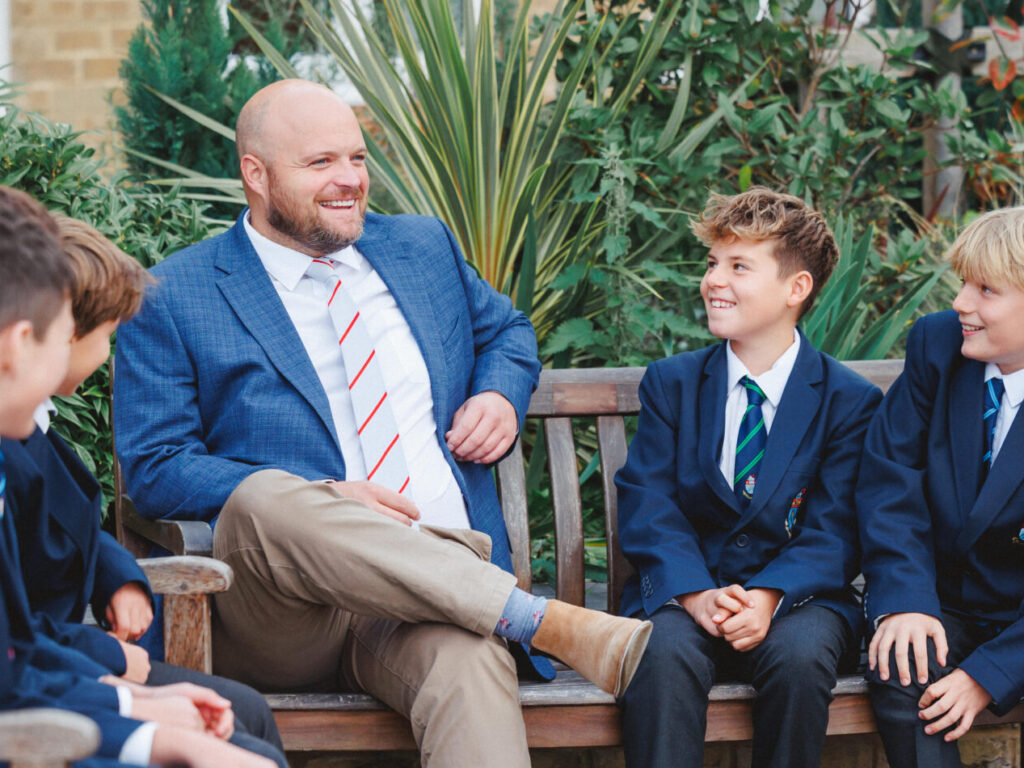 Man in blue blazer sits on bench with smiling schoolboys in uniforms outdoors, plants in background, relaxed conversation.