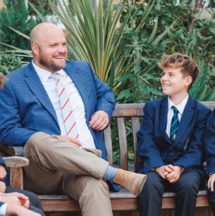 Man in blue blazer sits on bench with smiling schoolboys in uniforms outdoors, plants in background, relaxed conversation.