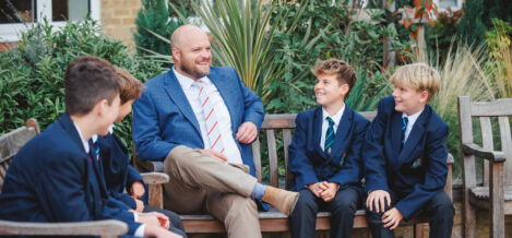Man in blue blazer sits on bench with smiling schoolboys in uniforms outdoors, plants in background, relaxed conversation.