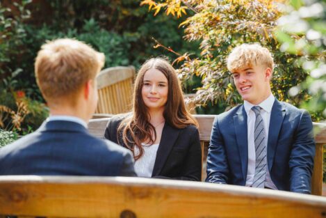 Three young professionals sit on a wooden bench outdoors, chatting in business attire with greenery in the background.