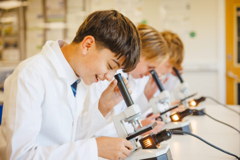 Students in white coats looking through microscopes in a science lab, focused on their slides under bright lamps