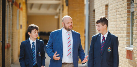 Teacher in a blue blazer walks with two students in uniforms along a brick building exterior.