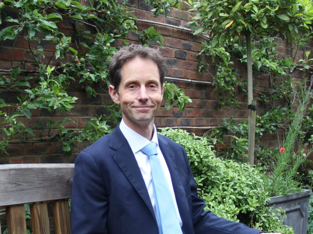Man in a navy suit and light blue tie sitting on a wooden bench in a lush garden with a brick wall behind him.