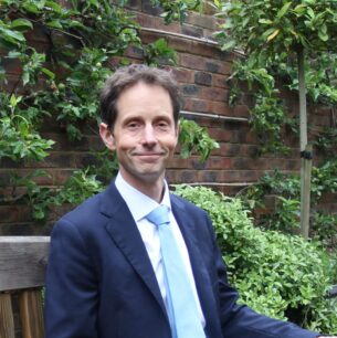 Man in a navy suit and light blue tie sitting on a wooden bench in a lush garden with a brick wall behind him.