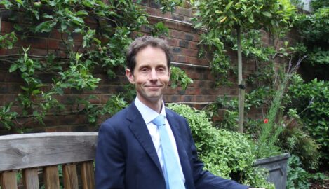 Man in a navy suit and light blue tie sitting on a wooden bench in a lush garden with a brick wall behind him.