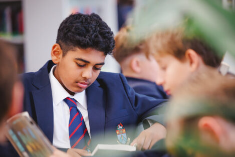 Boy in a navy blazer and striped tie reads a magazine at a table in a classroom setting.