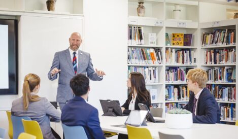 Male presenter in a gray suit speaks to a group in a bright library classroom with tall shelves behind him.
