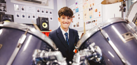 Smiling student in a blazer and tie sits among large drum kits in a classroom music room.