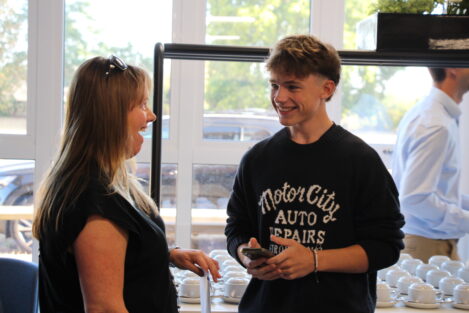 Two people smiling and talking near a row of white coffee cups on a counter by bright windows.