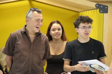 Three people standing in front of a bright yellow wall: a man on the left laughs, a smiling woman in the middle, and a young man on the right wearing glasses and holding papers.