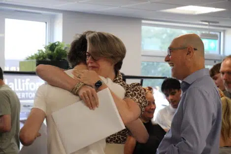 Two people hug warmly in a busy indoor event, smiling as they share a moment while others watch in the background.