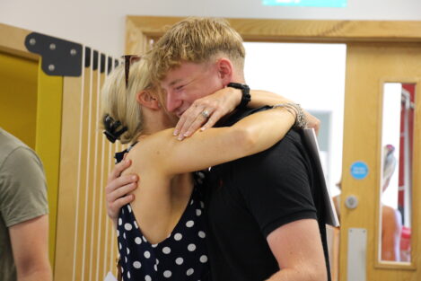 Two people hug warmly in a hallway, smiling with their faces pressed together; woman in a polka-dot dress and man in a black shirt.
