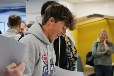 Smiling young man in a gray hoodie looking down at a table during a casual group event in an indoor setting.