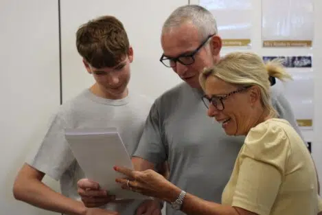 Three people lean in to read papers together, smiling in a bright room, likely colleagues or family members.